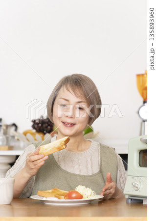 キッチンでトーストを焼いて朝食を食べるシニア女性 キッチンでトーストを焼いて朝食を食べるシニア女性 135893960