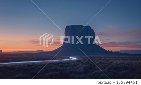 As the sun sets behind a massive desert monolith cars can be seen speeding by leaving behind a trail of shimmering blue light 135894451