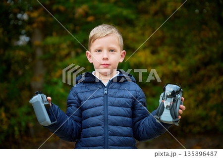 A young boy stands confidently outdoors, holding a camera in one hand and a small drone in the other, surrounded by colorful autumn leaves. 135896487