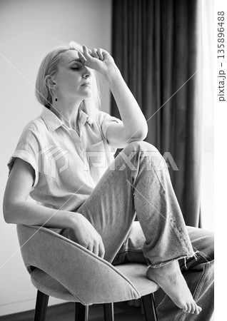 Woman seated on chair, in moment of distress or deep thought. Person with eyes closed and hand raised to their forehead, she feeling overwhelmed, stressed. Black and white image. 135896488