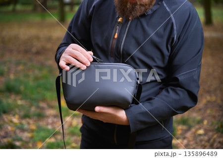 A man stands in a peaceful park, cradling a stylish bag close to his chest. Autumn leaves surround him as he enjoys the serene atmosphere of the afternoon. 135896489