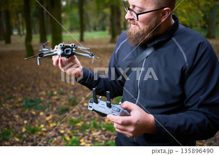 A bearded man in a black jacket holds a drone and controller in a colorful forest. Bright leaves surround him as he prepares for an exciting flight. 135896490