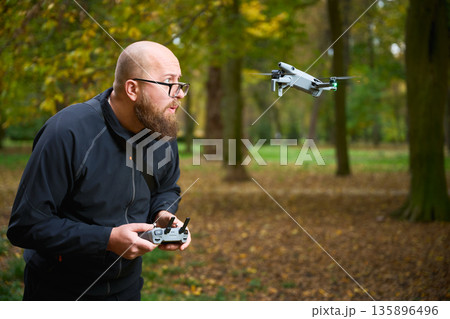 A person with a beard controls a flying drone in a vibrant park filled with colorful autumn leaves. The setting is peaceful and full of nature. A person with a beard controls a flying drone in a vibrant park filled with colorful autumn leaves. The setting is peaceful and full of nature. 135896496