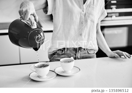 Cropped picture of woman pouring water into cup. Young female making tea for two. Woman holding kettle over two cups. Black and white image. 135896512