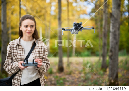 In a vibrant autumn forest, a young woman controls a drone with enthusiasm while surrounded by golden leaves and tall trees. 135896519