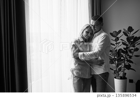 Happy couple hugging on light background at home. Pretty woman smiling and hugging her husband with closed eyes. Sweet couple in casual clothes. Black and white image. 135896534