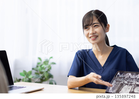 レントゲン画像が表示されたタブレットを手に患者と話す医療用スクラブを着た若い女性 135897248