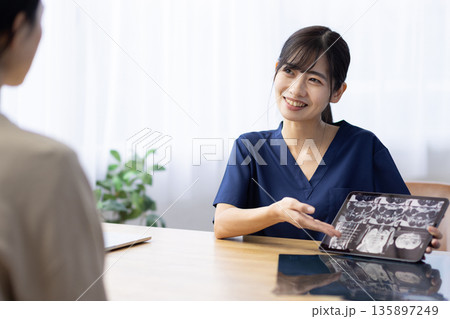 レントゲン画像が表示されたタブレットを手に患者と話す医療用スクラブを着た若い女性 135897249