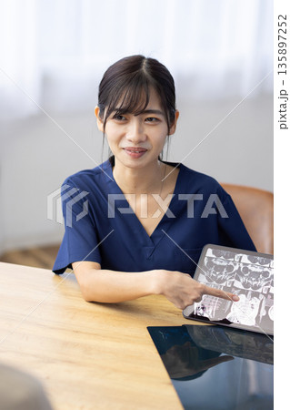 レントゲン画像が表示されたタブレットを手に患者と話す医療用スクラブを着た若い女性 レントゲン画像が表示されたタブレットを手に患者と話す医療用スクラブを着た若い女性 135897252