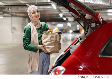 A woman wearing a hijab smiles while holding a bag of groceries near the trunk of a red car in a parking garage 135897298