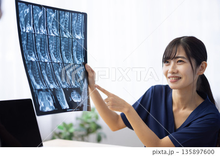 レントゲン写真を見せながら患者に説明する紺色の医療用スクラブを着た若い日本人女性 135897600