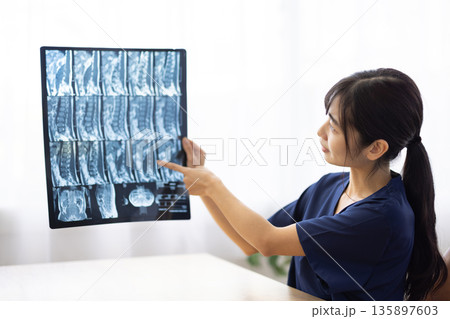 レントゲン写真を見せながら患者に説明する紺色の医療用スクラブを着た若い日本人女性 135897603