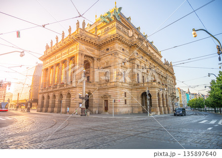 A historic National theater building stands under the morning sunlight. The street is empty, and trams run along the tracks. Trees are visible, creating a city scene at dawn. A historic National theater building stands under the morning sunlight. The street is empty, and trams run along the tracks. Trees are visible, creating a city scene at dawn. 135897640