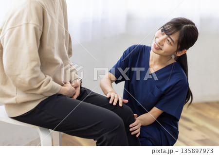 患者の膝に手を当てて笑顔で話す紺色のスクラブを着た若い女性 患者の膝に手を当てて笑顔で話す紺色のスクラブを着た若い女性 135897973