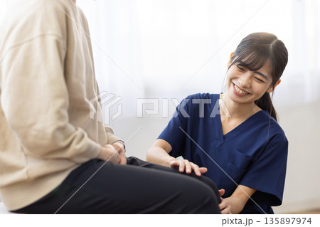患者の膝に手を当てて笑顔で話す紺色のスクラブを着た若い女性 患者の膝に手を当てて笑顔で話す紺色のスクラブを着た若い女性 135897974
