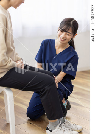 患者の膝に手を当てて笑顔で話す紺色のスクラブを着た若い女性 135897977