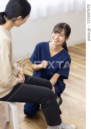 患者の膝に手を当てて笑顔で話す紺色のスクラブを着た若い女性 135897978