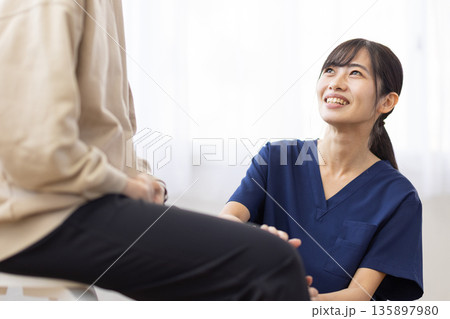 患者の膝に手を当てて笑顔で話す紺色のスクラブを着た若い女性 135897980