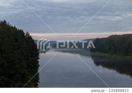 a wide river and trees on the shore during sunset with a multicolored sky, a landscape on the river at dusk with a multicolored sky that is reflected in the water of the river 135898923