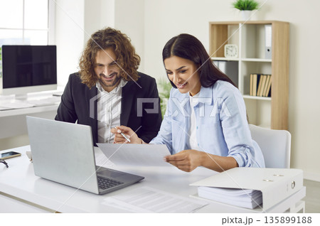 Two business accountants sitting at an office desk together, doing paperwork and using a laptop 135899188