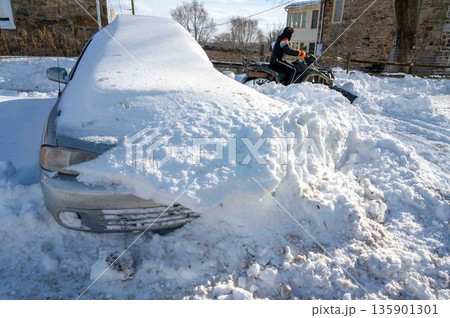 Man on ATV plows snow clearing driveway with a car buried in snowdrift Man on ATV plows snow clearing driveway with a car buried in snowdrift 135901301