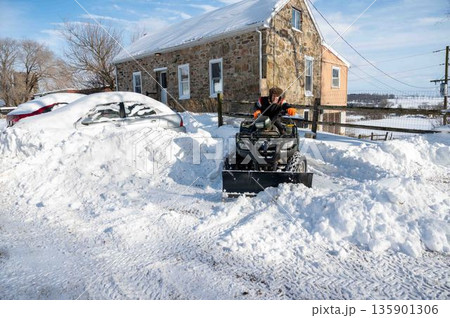 Man on ATV plows snow clearing driveway with a car buried in snowdrift Man on ATV plows snow clearing driveway with a car buried in snowdrift 135901306