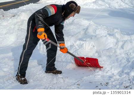 A man in a snowsuit and fur hat shovels snow on a rural driveway with street A man in a snowsuit and fur hat shovels snow on a rural driveway with street 135901314