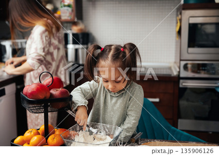 Child mixes ingredients while another person prepares food in a kitchen during a busy afternoon with fruit and cooking supplies around Child mixes ingredients while another person prepares food in a kitchen during a busy afternoon with fruit and cooking supplies around 135906126