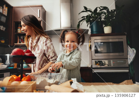 Mom and daughter bake together in a kitchen filled with fruits and cooking tools during daytime, creating sweet memories with smiles and laughter 135906127