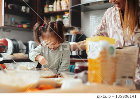 Child and adult cooking together in a kitchen while making a dessert with flour, sugar, and other ingredients on the table during a weekend afternoon Child and adult cooking together in a kitchen while making a dessert with flour, sugar, and other ingredients on the table during a weekend afternoon 135906141
