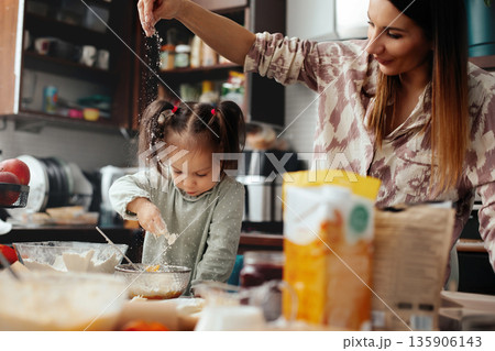 Mother and daughter cook together in the kitchen, making a dessert with flour and sugar on a busy countertop filled with cooking items during the afternoon 135906143
