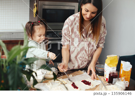 Family members prepare baked goods together in kitchen during afternoon hours with ingredients and tools on the table 135906146