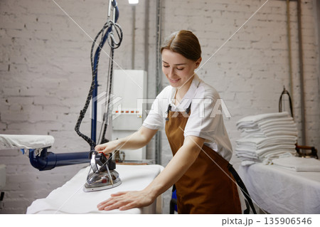A woman worker wearing an apron is skillfully ironing a shirt on a table A woman worker wearing an apron is skillfully ironing a shirt on a table 135906546