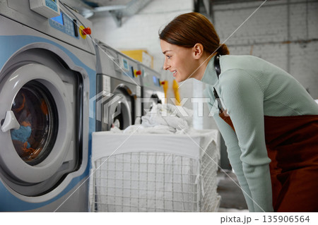 A woman worker is looking at a washing machine inside a laundromat A woman worker is looking at a washing machine inside a laundromat 135906564