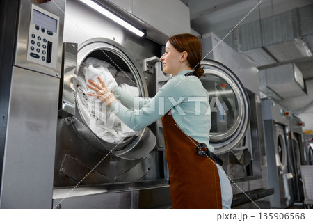 A woman worker is loading various clothes into a washing machine 135906568