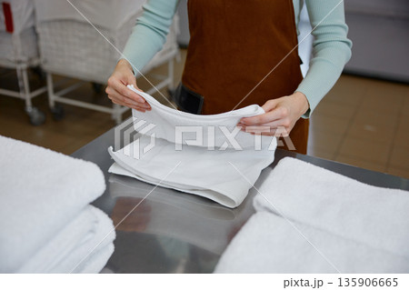 Female worker is carefully folding clean towels in a bustling laundromat Female worker is carefully folding clean towels in a bustling laundromat 135906665