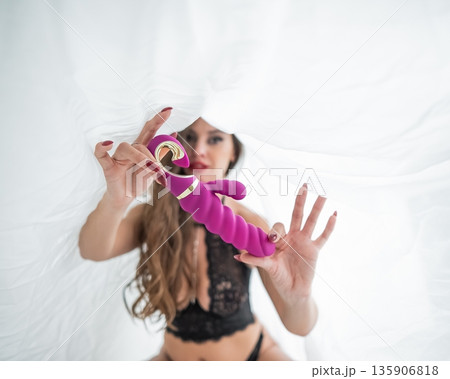 A woman in lingerie sits under a sheet and holds a vibrator. A woman in lingerie sits under a sheet and holds a vibrator. 135906818