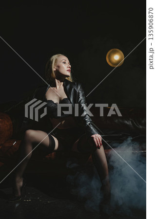 A Caucasian woman smokes from a mouthpiece while sitting on a leather sofa in her underwear. 135906870