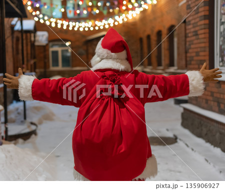 Rear view of santa claus carrying sack of gifts along decorated street. Rear view of santa claus carrying sack of gifts along decorated street. 135906927