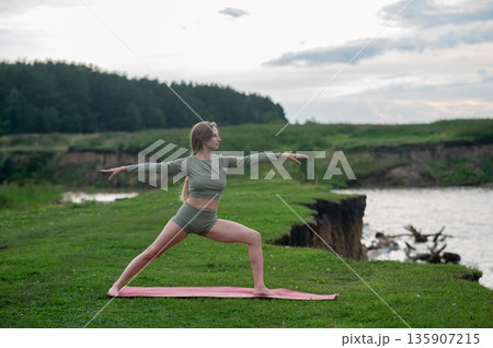Caucasian woman doing yoga on the river bank. Warrior pose. 135907215