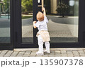 Curious baby standing outdoors and touching for door handle. Back view Cute child opening door, exploring, modern childhood lifestyle, safety and curiosity concept in urban city 135907378