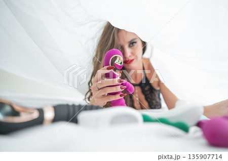 Woman in lace lingerie holds a dildo. A beautiful Caucasian woman lies under the sheets and holds a dildo. Woman in lace lingerie holds a dildo. A beautiful Caucasian woman lies under the sheets and holds a dildo. 135907714