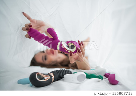 Woman in lace lingerie holds a dildo. A beautiful Caucasian woman lies under the sheets and holds a dildo.  135907715