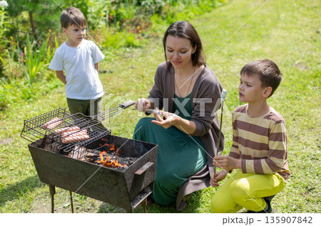 Caucasian woman grilling sausages on a barbecue with her two sons. Caucasian woman grilling sausages on a barbecue with her two sons. 135907842