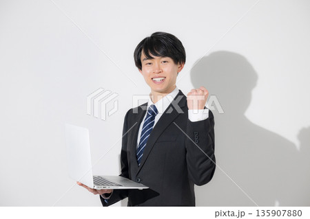 白背景でパソコンを持つスーツの若い大学生のような男性　新卒や就職のビジネス就活の採用　ガッツポーズ 135907880