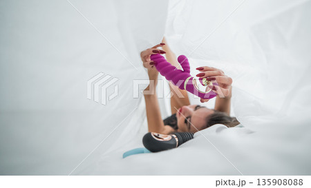 Woman in lace lingerie holds a dildo. A beautiful Caucasian woman lies under the sheets and holds a dildo.  135908088