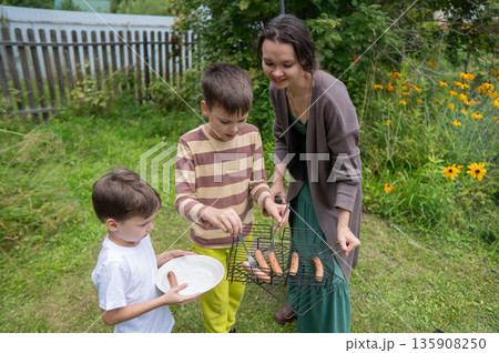 Caucasian woman grilling sausages on a barbecue with her two sons. Caucasian woman grilling sausages on a barbecue with her two sons. 135908250