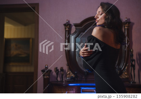A beautiful brunette in a black evening dress poses in front of a retro-style mirror. A beautiful brunette in a black evening dress poses in front of a retro-style mirror. 135908282