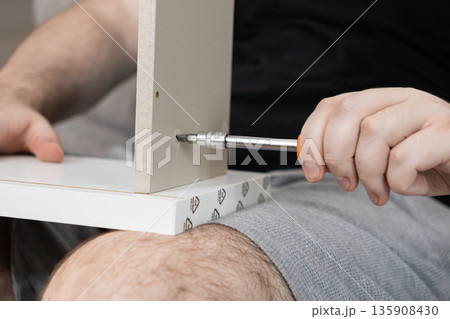 Male hand uses a screwdriver to tighten a screw into a furniture panel, highlighting home assembly, do-it-yourself tasks, and practical residential improvement Male hand uses a screwdriver to tighten a screw into a furniture panel, highlighting home assembly, do-it-yourself tasks, and practical residential improvement 135908430