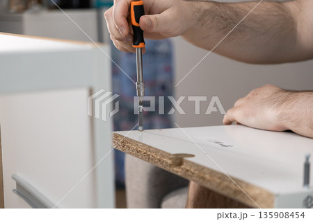 A person's hands expertly fasten a screw into white particle board using an orange screwdriver, embodying home improvement, practical DIY, and furniture assembly projects 135908454